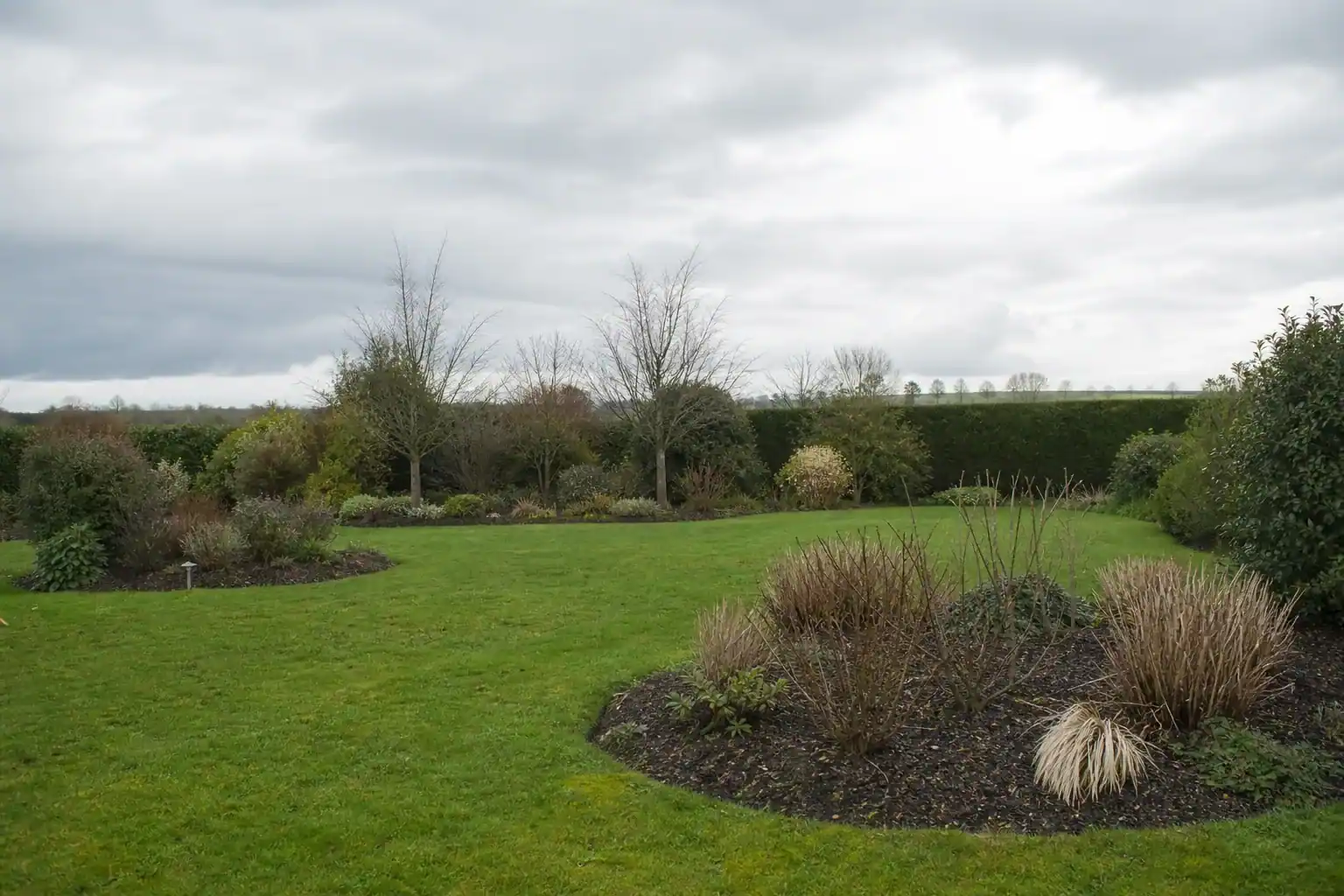 Jardin de campagne dégagé à Cerisé protégé par une haie brise-vent, pelouse structurée et massifs abrités, grand ciel ouvert typique de la plaine près de l'aérodrome.