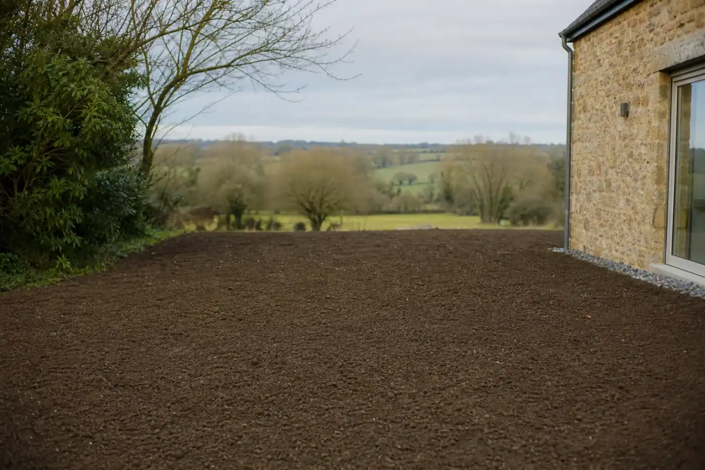 Terrain fraîchement nivelé et prêt pour un semis de gazon dans un jardin pavillonnaire du secteur d'Alençon, terre végétale sombre bien affinée, bocage normand flou en arrière-plan, lumière douce de fin d'après-midi.