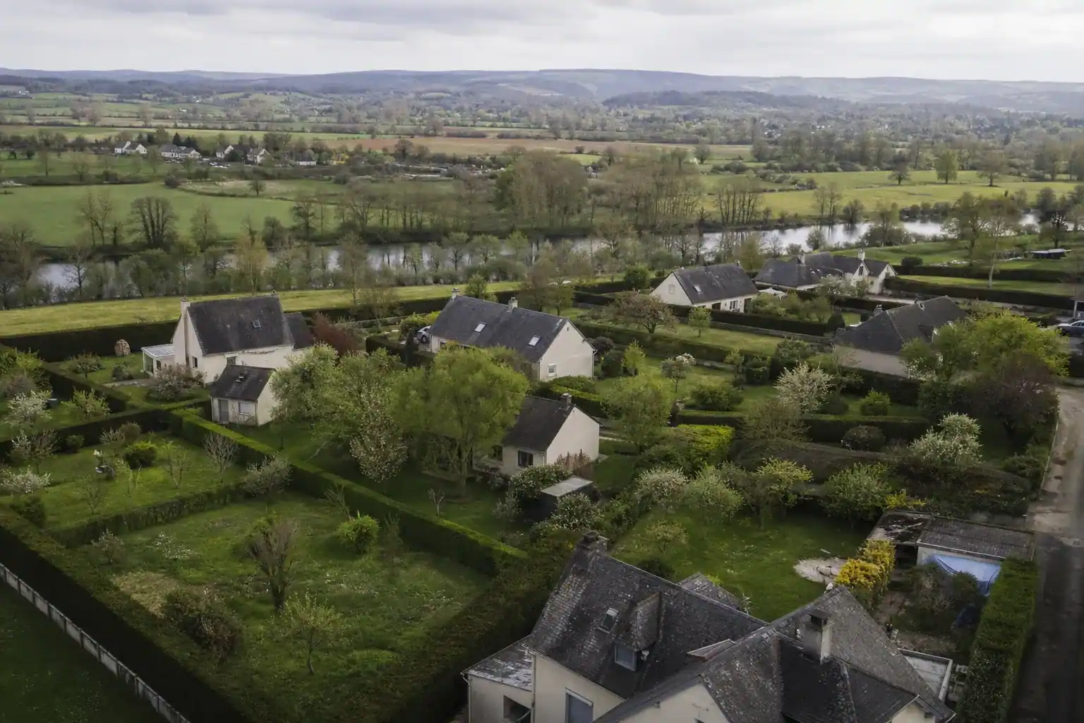 Vue aérienne basse d'un quartier pavillonnaire de la couronne d'Alençon : jardins verts contigus, haies taillées de bocage, la vallée de la Sarthe en arrière-plan, ciel normand doux de fin d'après-midi.