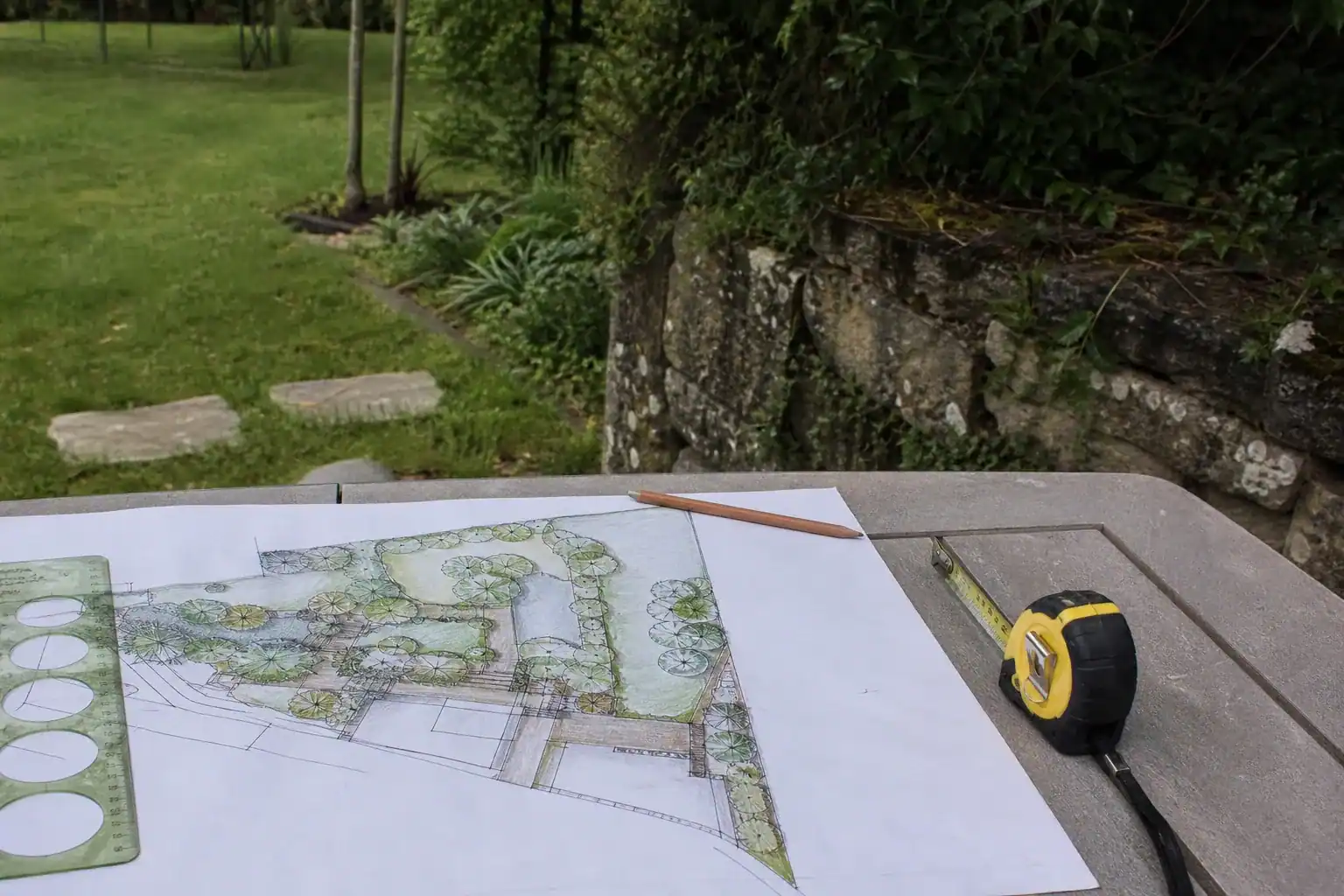Plan de jardin dessiné à la main posé sur une table d'extérieur, crayon et mètre ruban, coin de pelouse réelle et muret de granit visibles derrière.