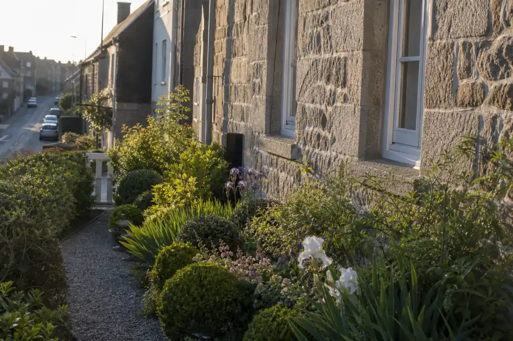 Jardin de devant étroit d'une maison ouvrière en granite le long d'un village-rue de Damigny, massif net et allée soignée, lumière rasante de fin de journée.