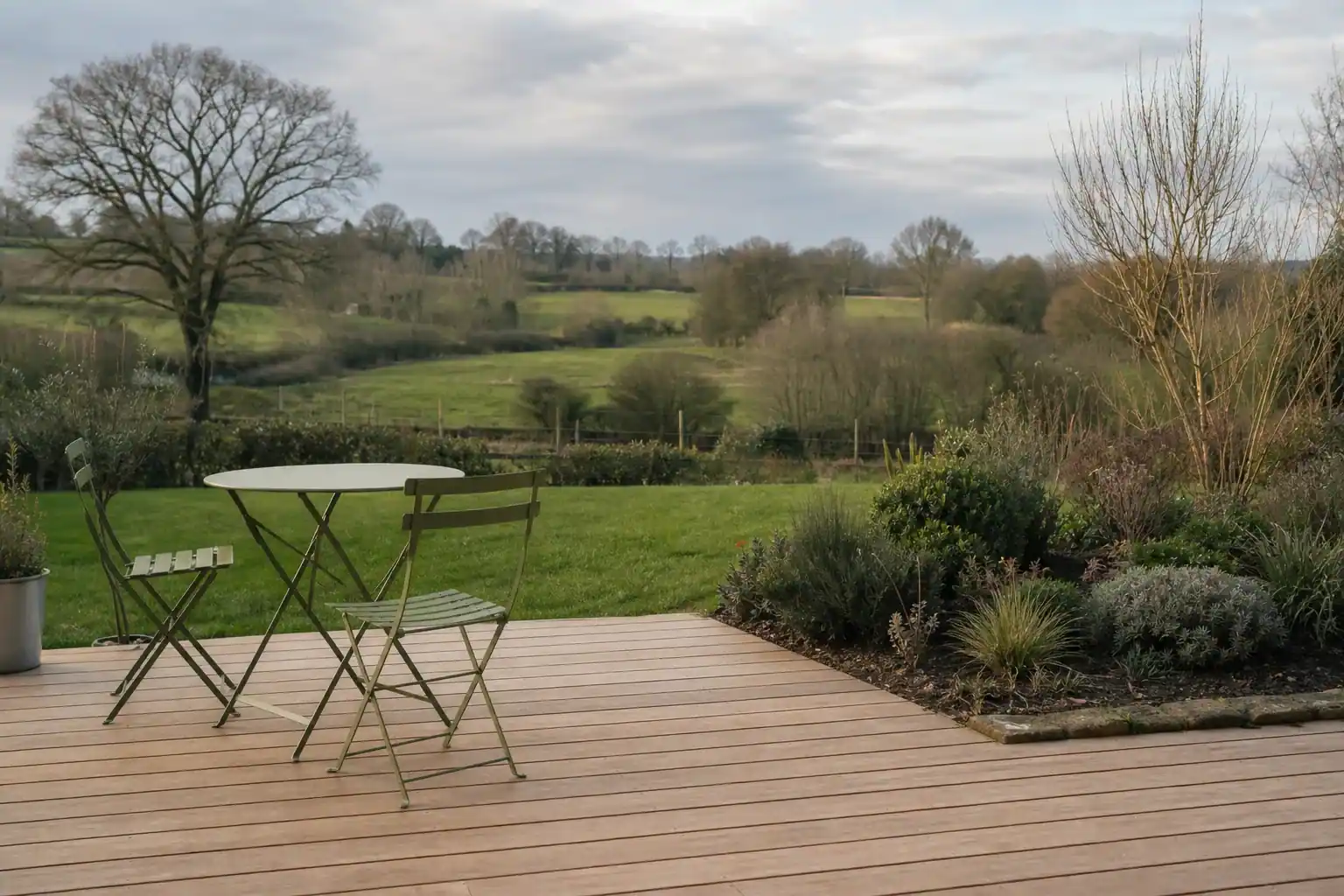 Terrasse en bois fraîchement posée ouvrant sur une pelouse d'un jardin du secteur d'Alençon, lames alignées au cordeau, massif en bordure, table et deux chaises simples, lumière de fin d'après-midi, bocage normand en fond.